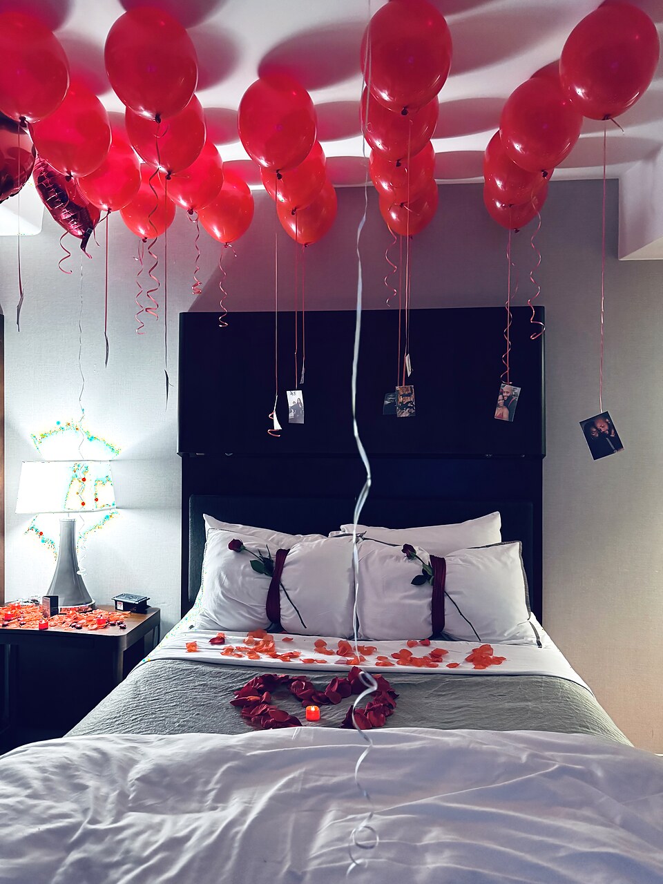 Anniversary hotel bed styled with red balloons, roses, petals, and keepsake photos